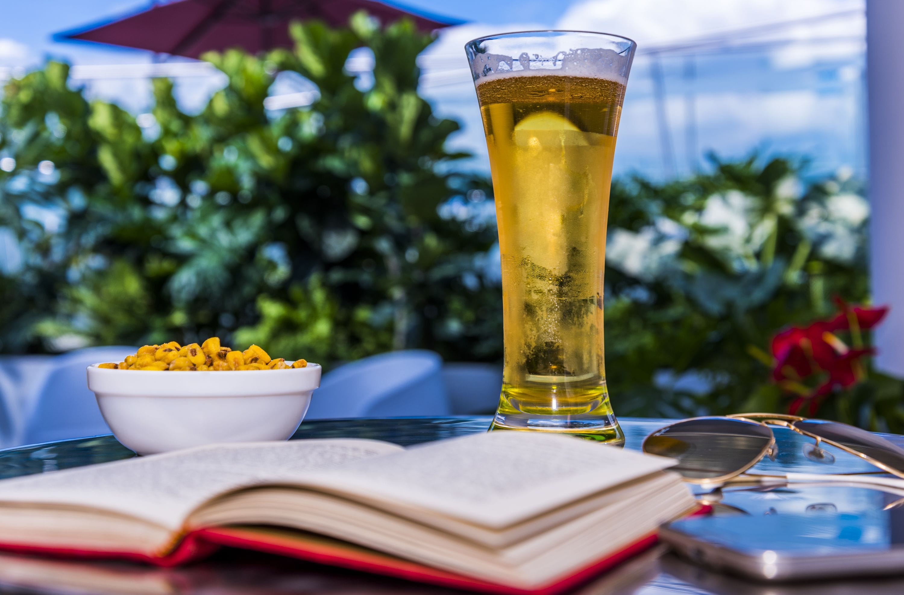 Una mesa con un vaso alto de cerveza, un bol de aperitivos, un libro abierto y un par de lentes.