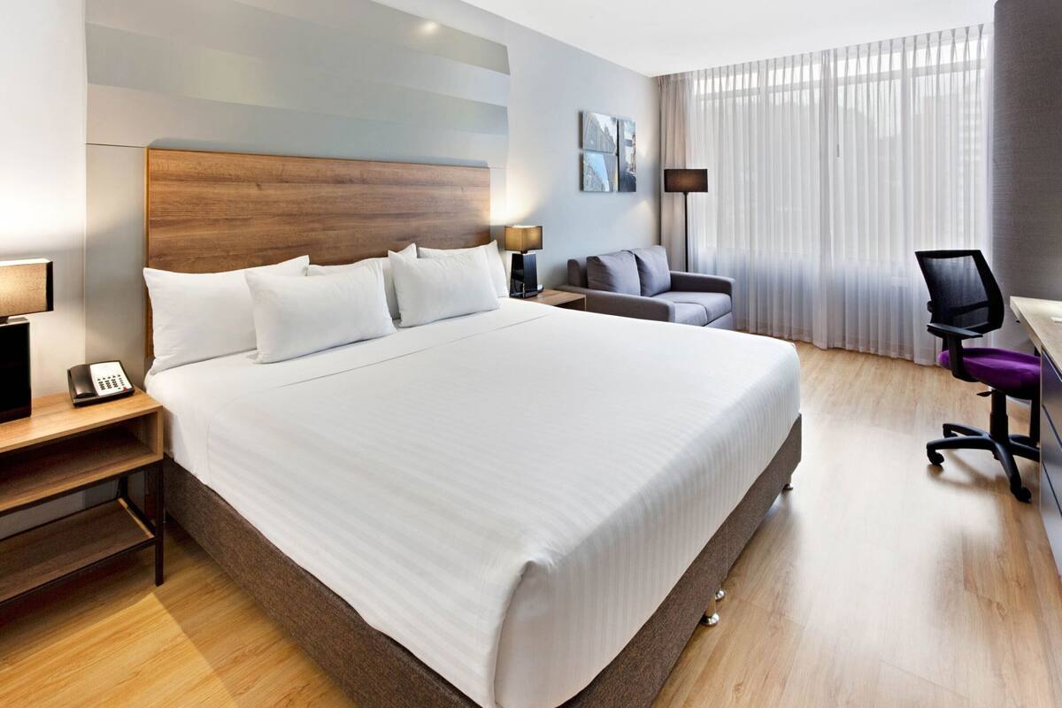 A hotel room with a large bed, bedside tables, a desk with a chair, a lamp, and a window with curtains.