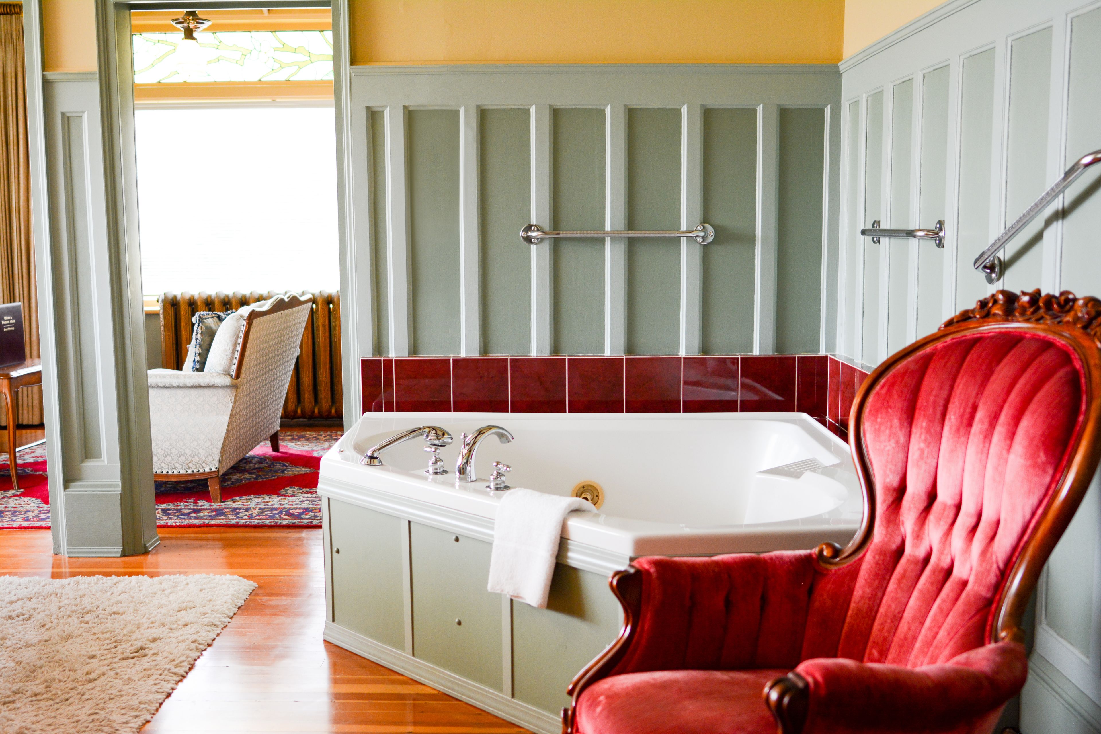 A bathroom with a bathtub, a red chair, and a window with curtains.