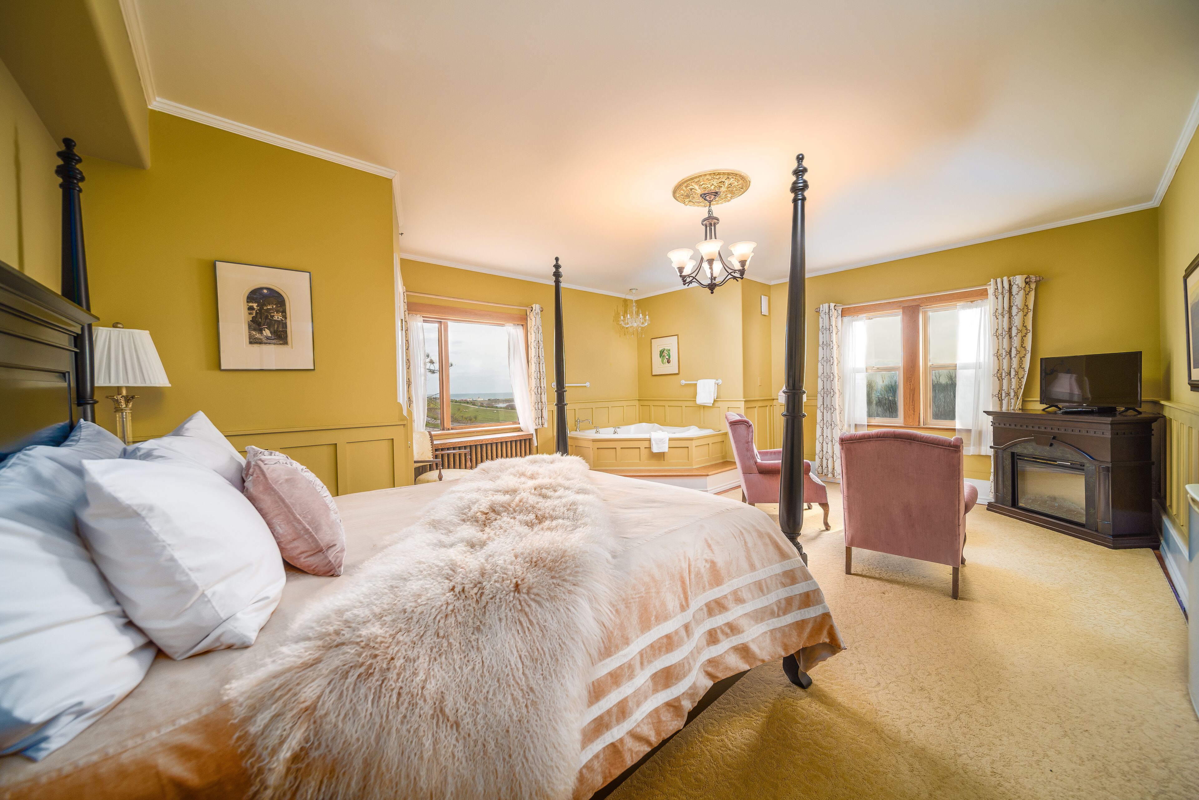 A bedroom with a four-poster bed, a sitting area with a chair and a fireplace, and a view of the outdoors through a window.
