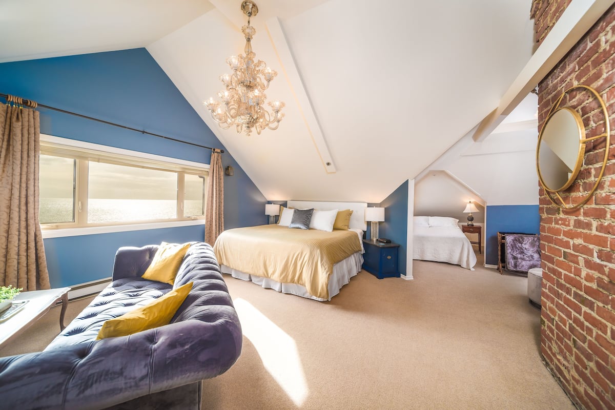 A bedroom with a large bed, a sofa, a chandelier, and a view through a window.