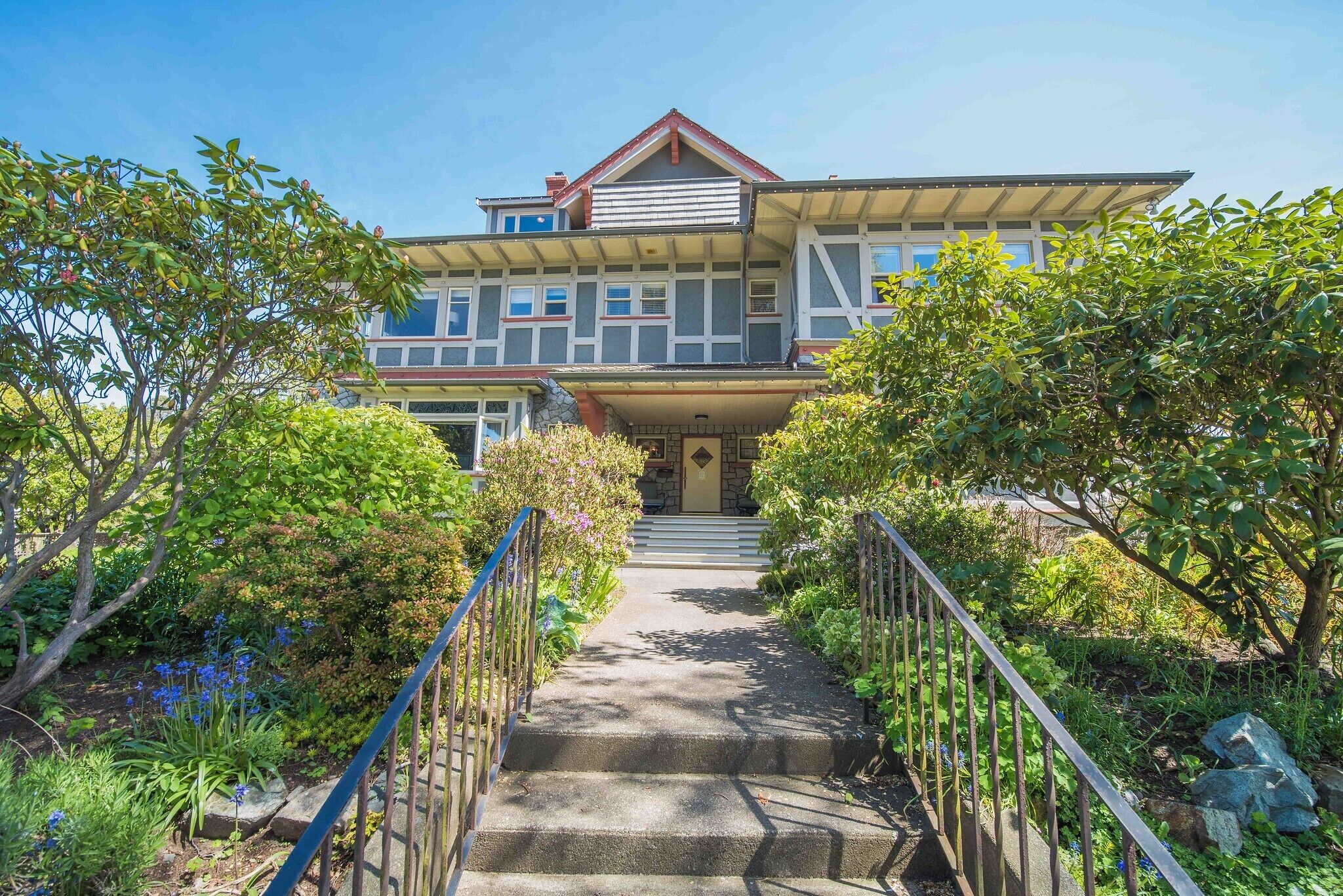 A two-story house with a front porch, a staircase leading to the entrance, and a garden with various plants and trees.