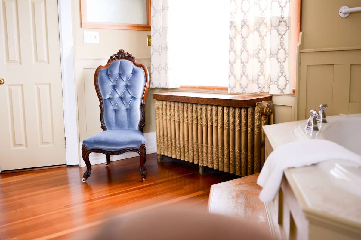 A room with a wooden floor, a blue upholstered chair, a radiator, and a white bathtub.