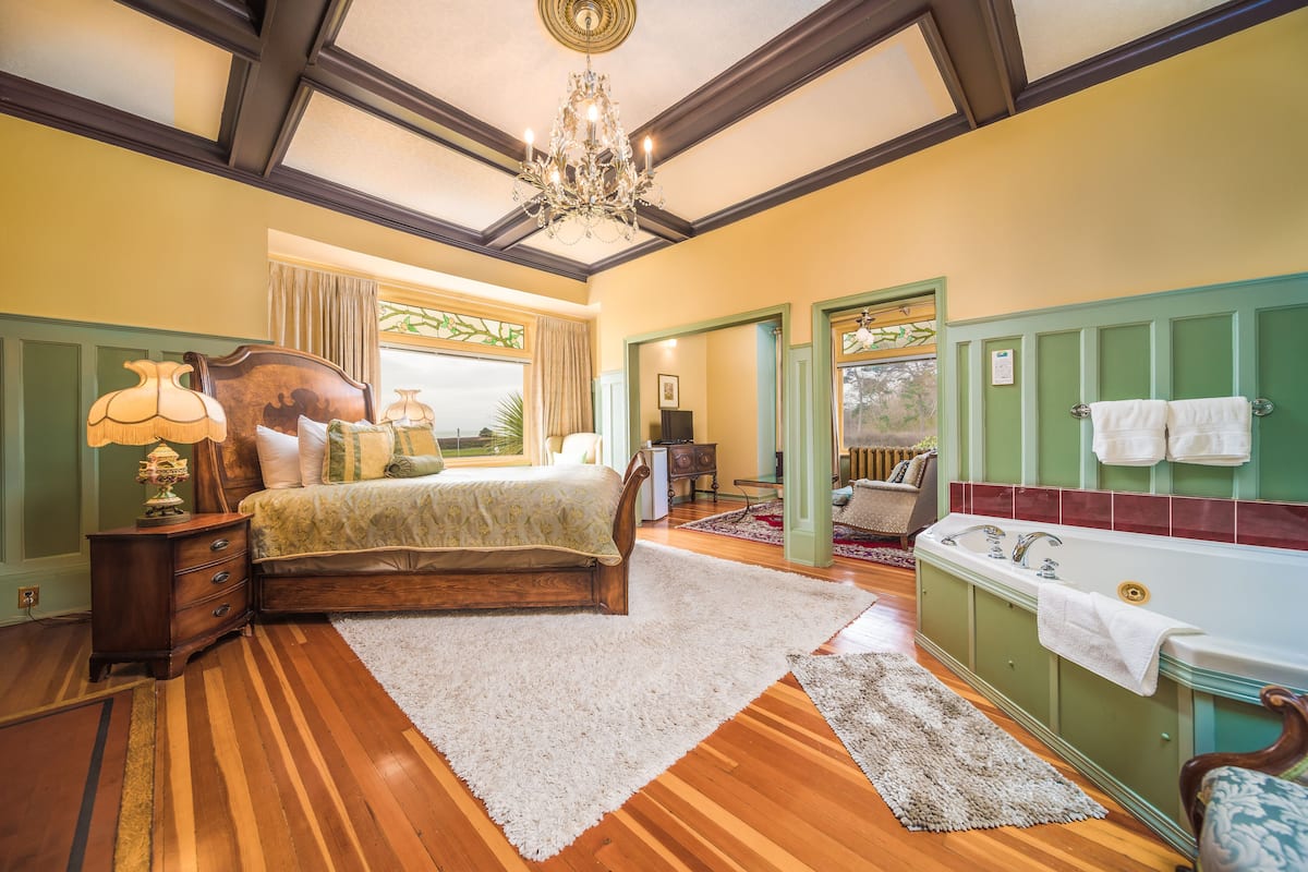 A bedroom with a wooden bed, a chandelier, a green vanity with a bathtub, and a large window with curtains.