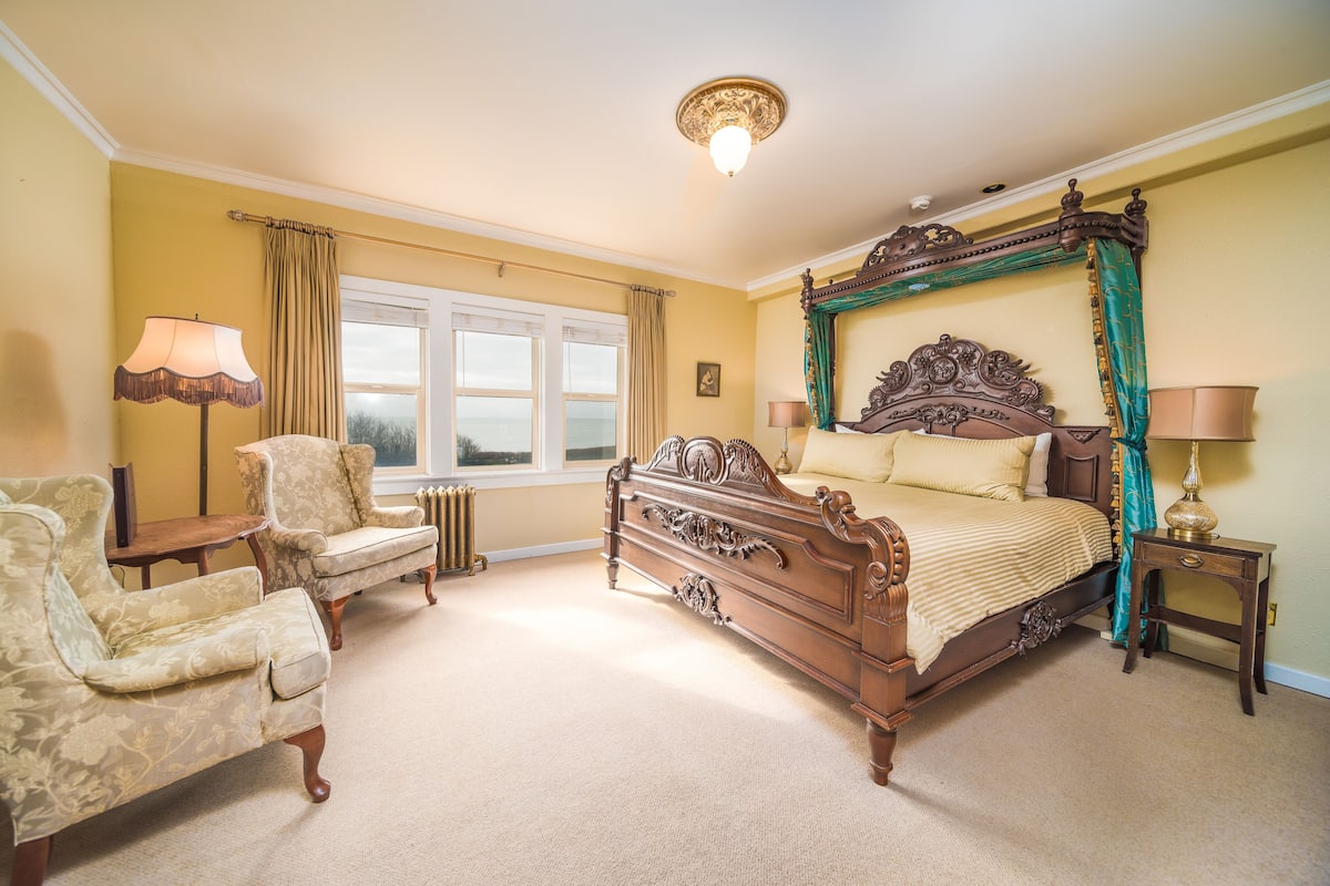 A bedroom with a four-poster bed, two armchairs, a table, and a window with curtains.