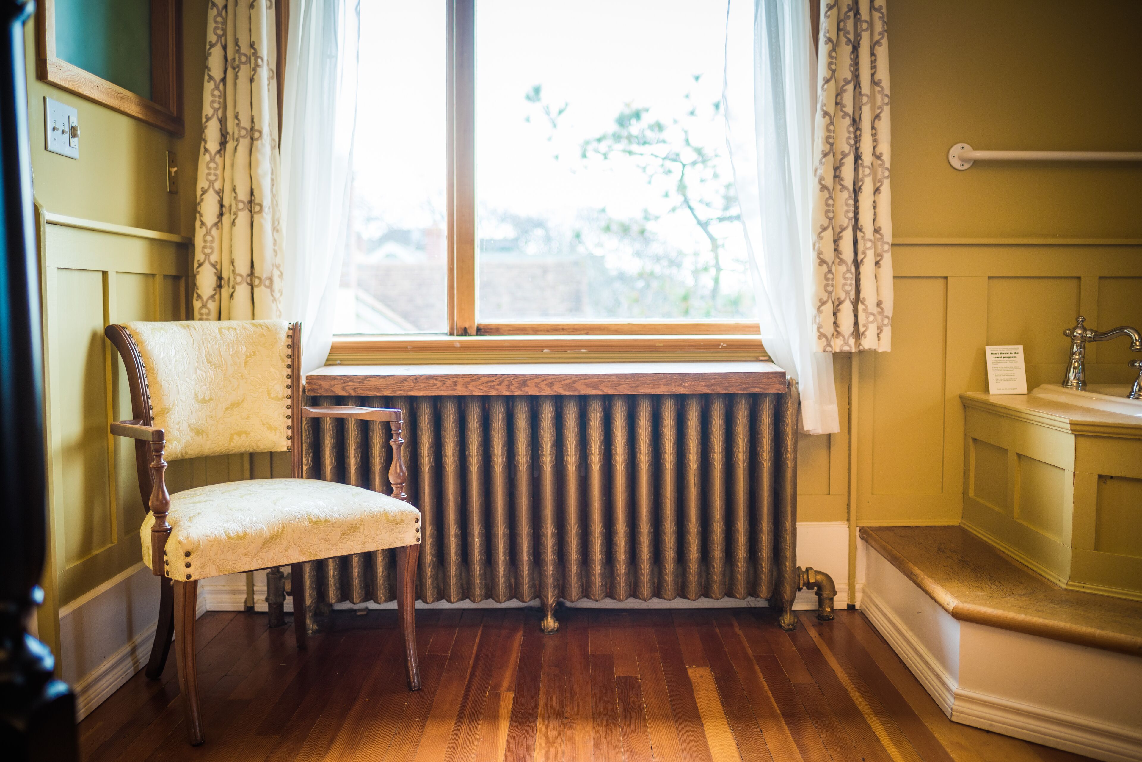 Um quarto com uma cadeira de madeira, um radiador e uma janela com cortinas.