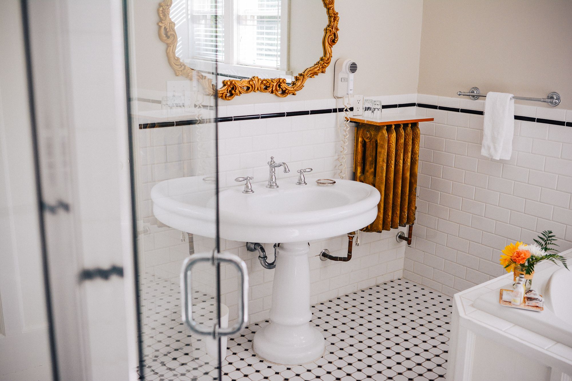A bathroom with a white pedestal sink, a gold ornate mirror, and a black and white tiled floor.