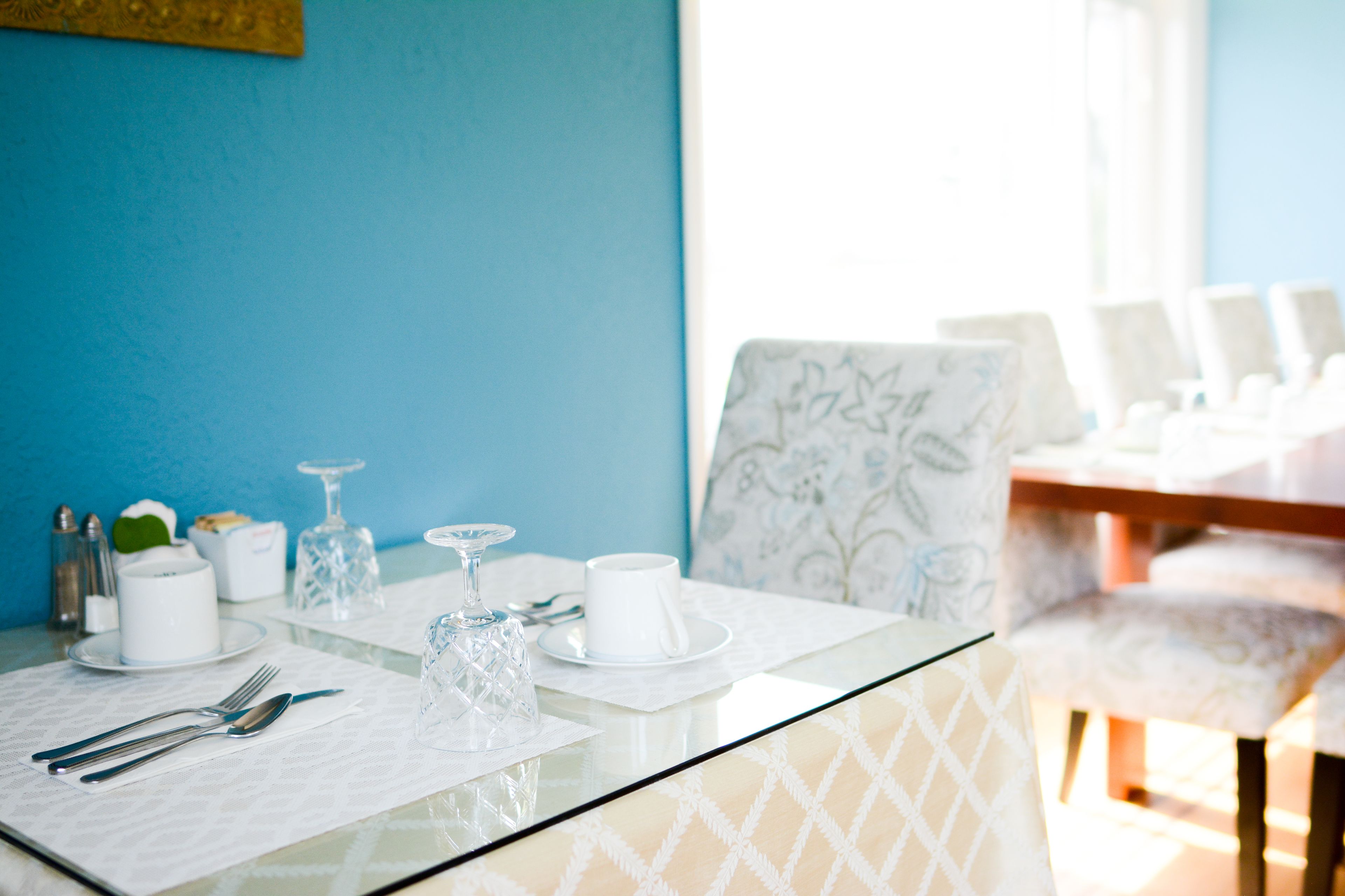 Una mesa de comedor dispuesta con vajilla blanca, una copa y un objeto decorativo sobre una mesa con cubierta de vidrio en una habitación con paredes azules y sillas estampadas.