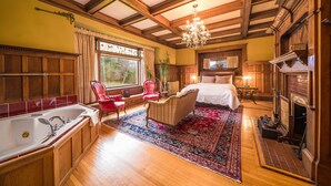 Una habitación amplia con una cama grande, una zona de estar con un sillón rojo, piso de madera, un ventanal y una chimenea.
