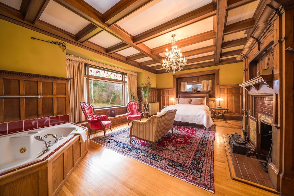 A spacious room with a large bed, a sitting area with a red chair, a wooden floor, a large window, and a fireplace.