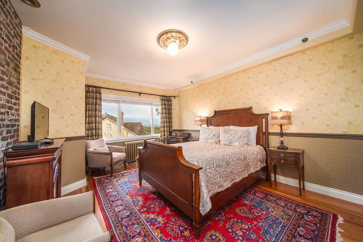 A bedroom with a large bed, a wooden dresser, a chair, a nightstand with a lamp, and a view of the outside.