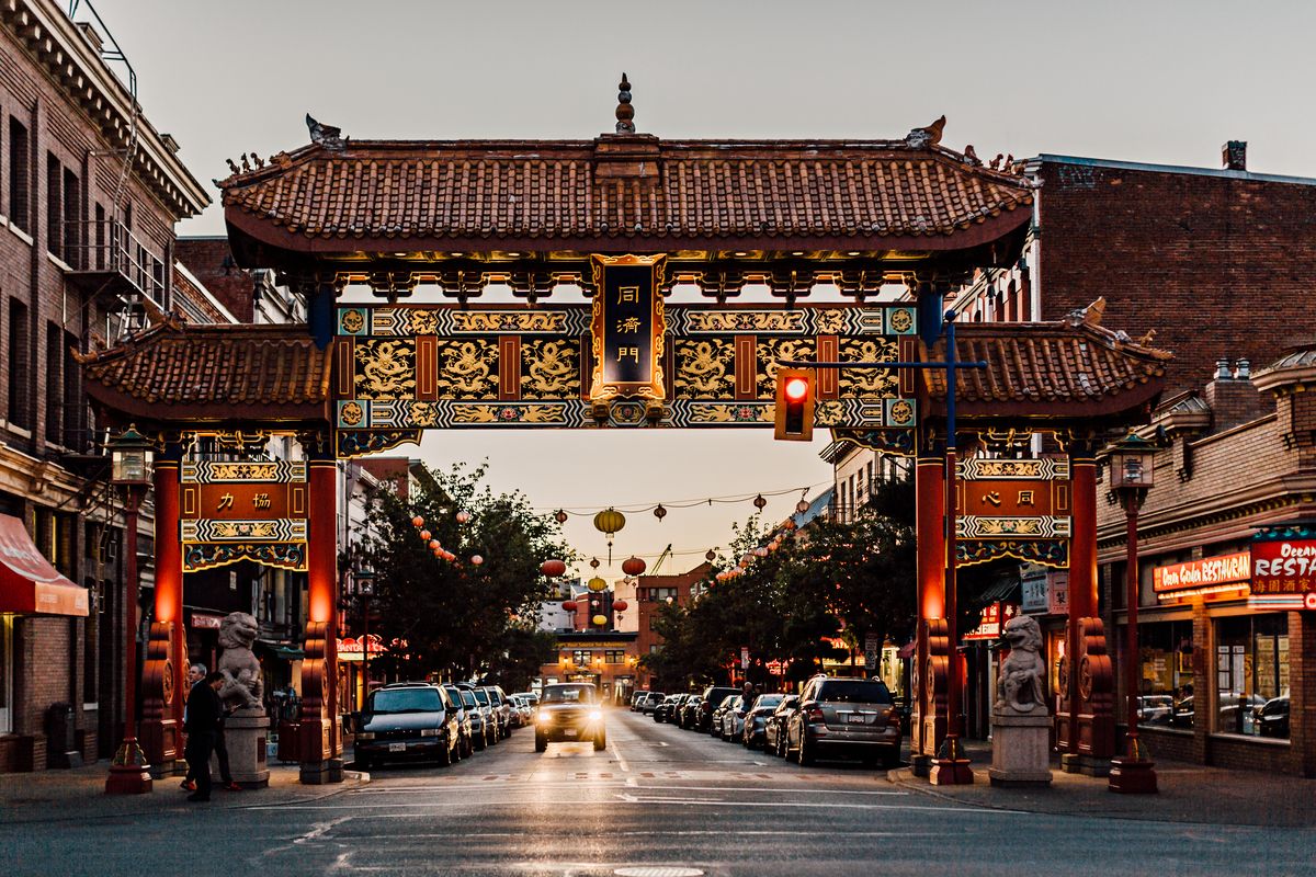 A traditional Chinese gate with intricate designs and characters, leading to a street lined with parked cars and buildings.