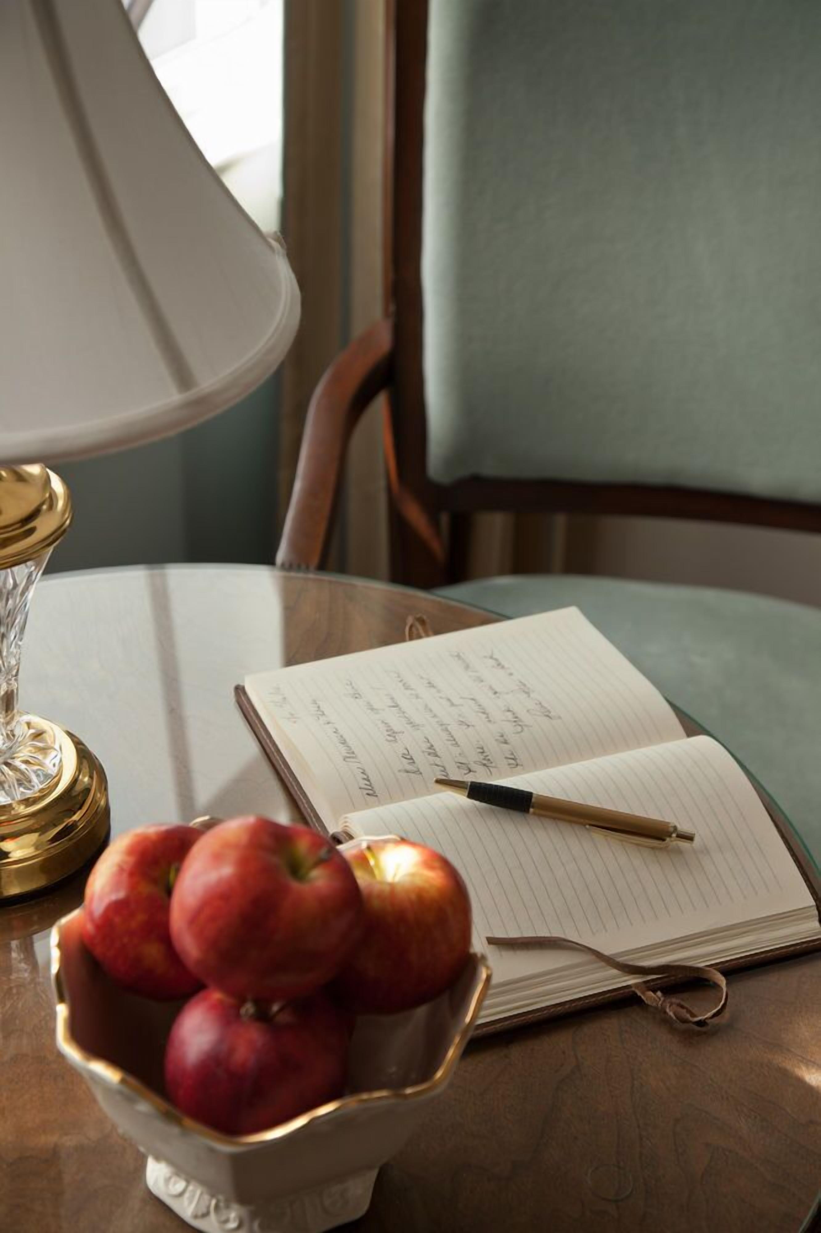 A table with a lamp, a bowl of apples, a notebook, and a pen.