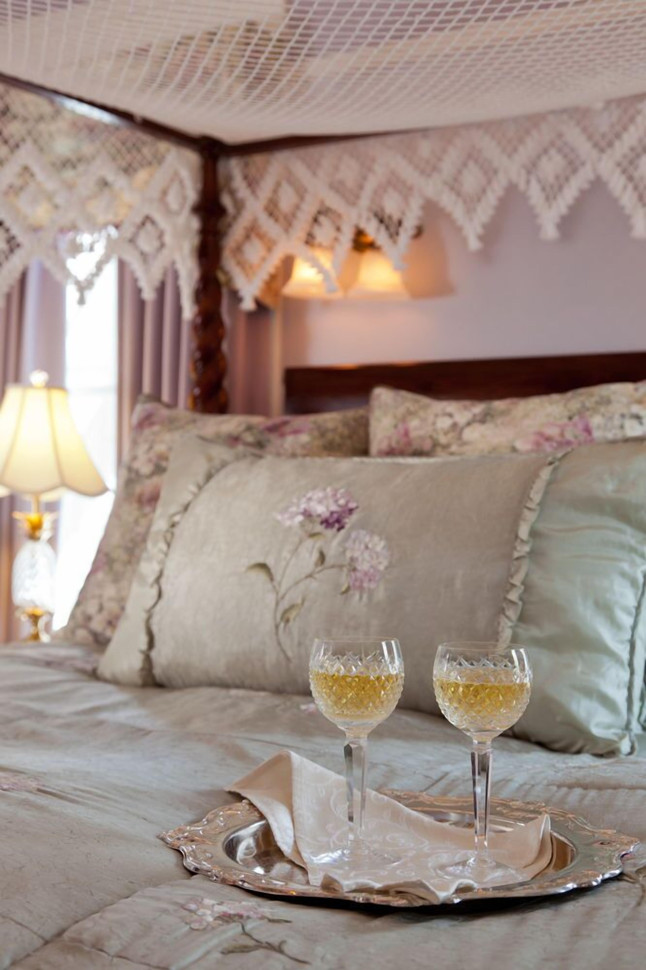 A bed with a floral-patterned bedspread, two glasses of white wine on a tray, and a lace headboard.