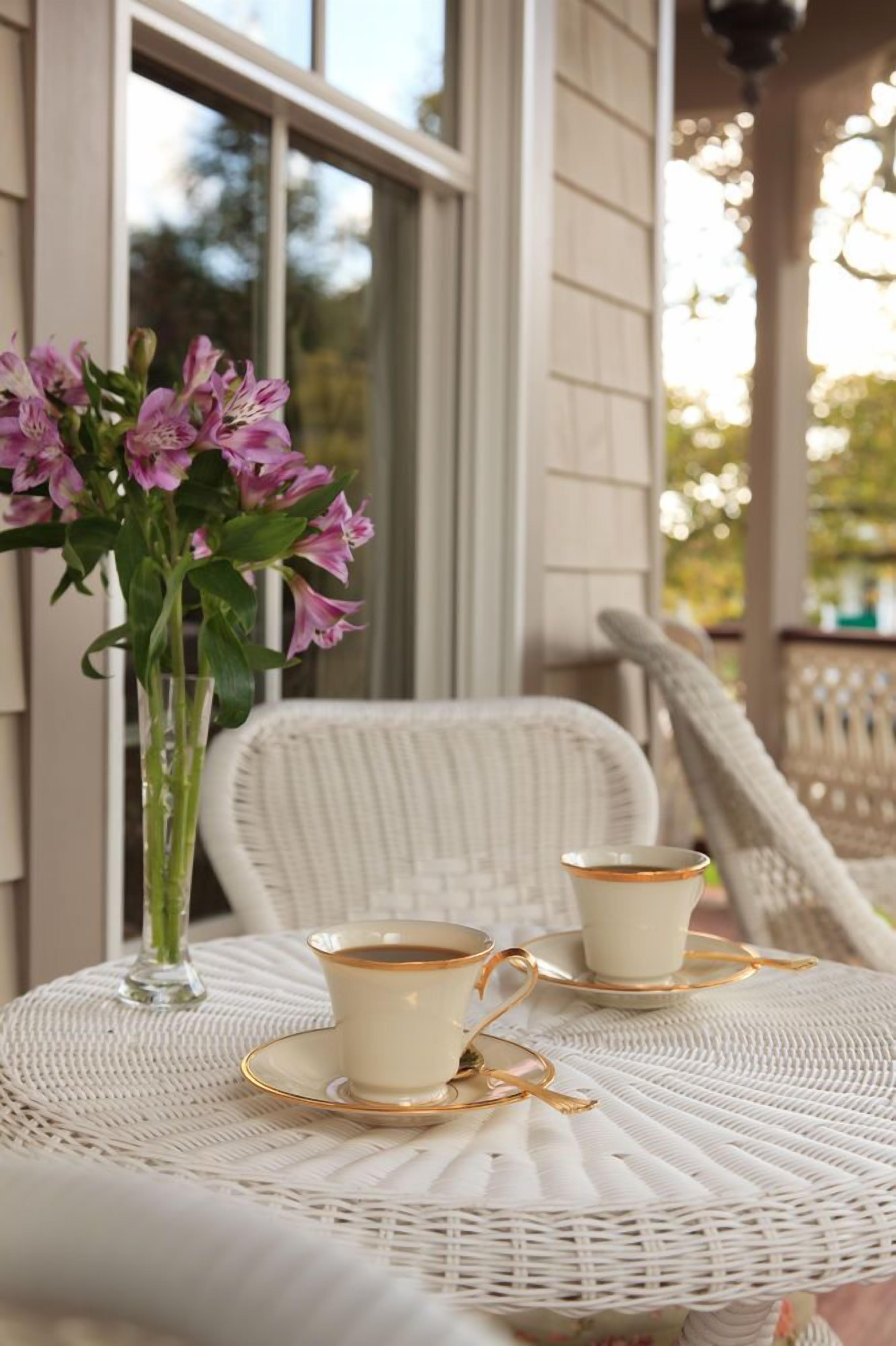 A wicker table with two white cups and saucers, a vase with pink flowers, and a wicker chair.