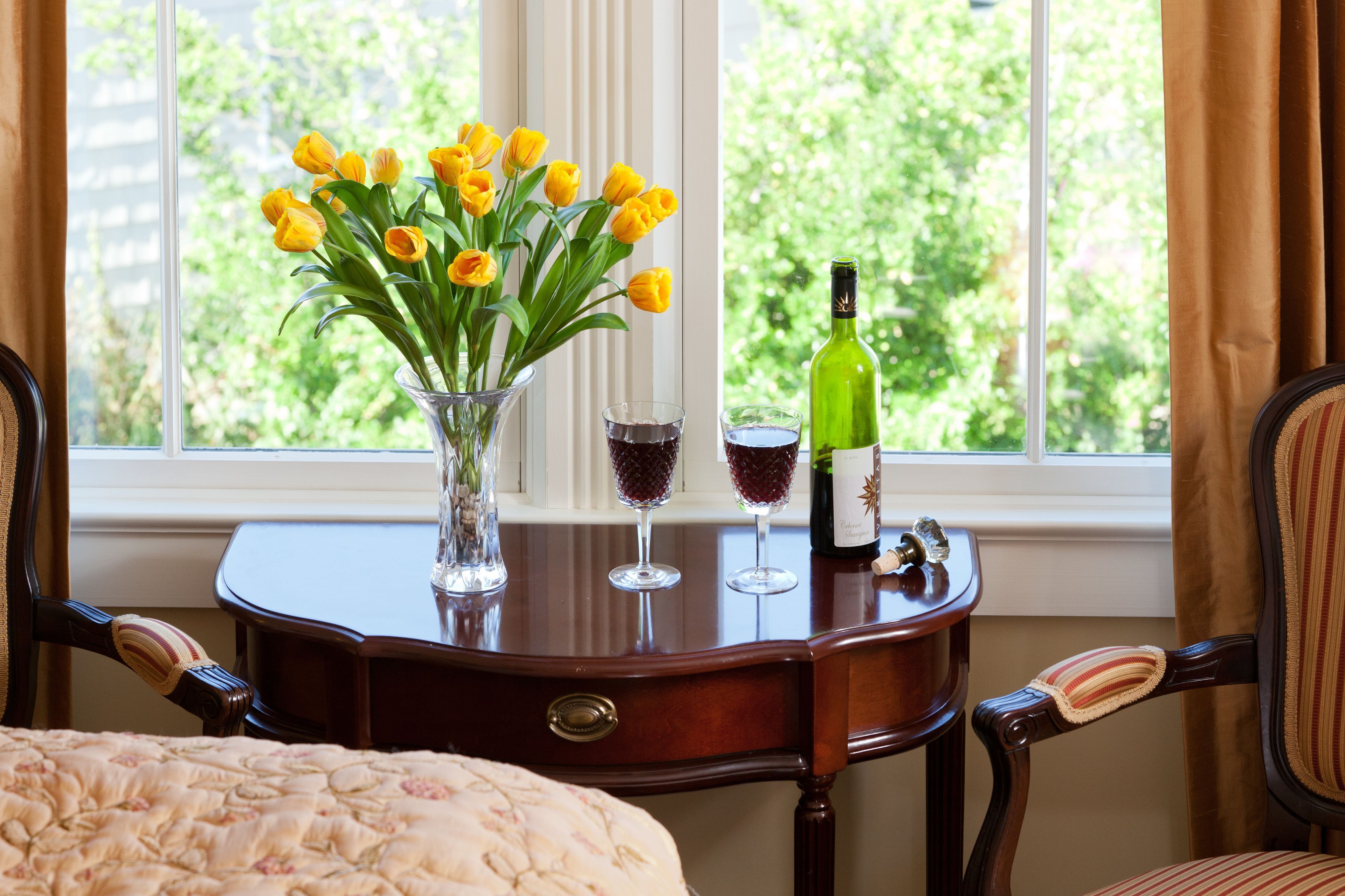 Ein Holztisch mit einer Vase voller gelber Tulpen, zwei Weingläsern und einer Weinflasche.