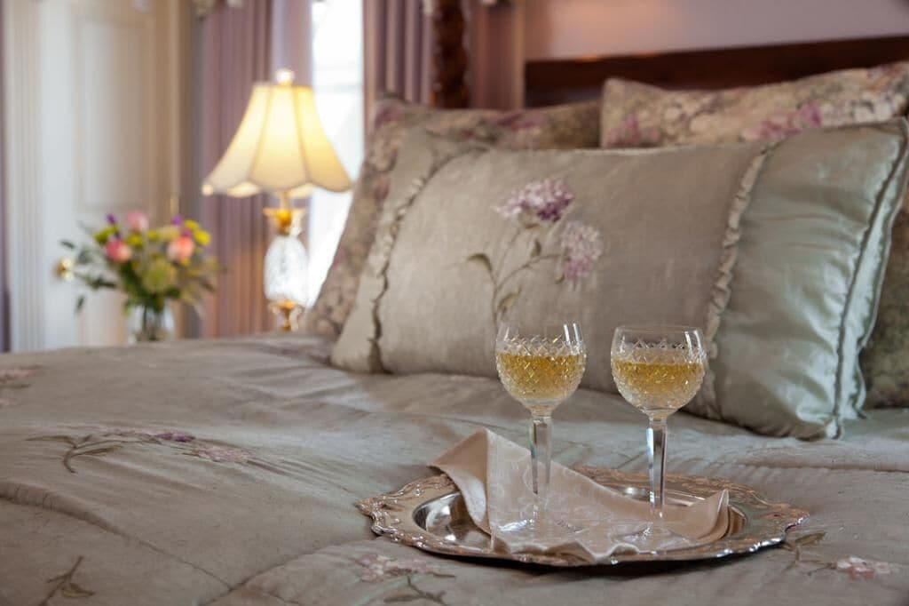 Una cama con ropa de cama floral, dos copas de vino blanco sobre una bandeja y una lámpara al fondo.