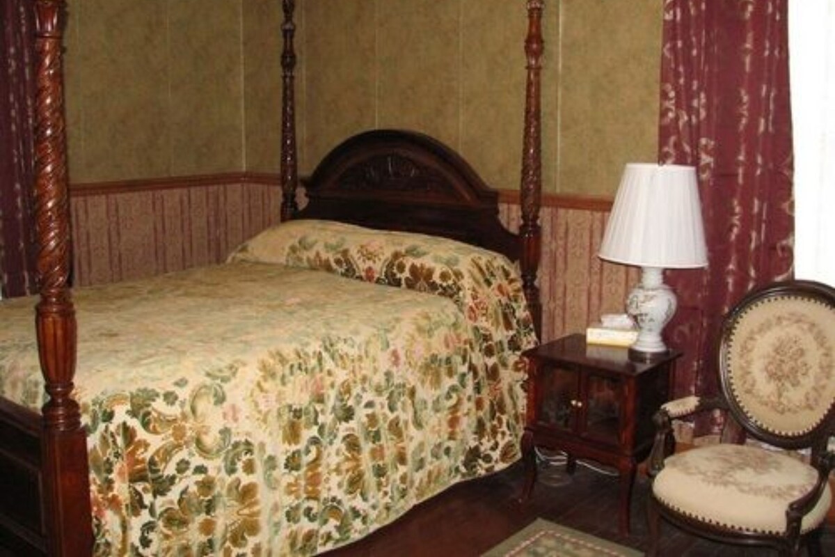 Een hemelbed met een bloemenprint, een nachtkastje met een lamp en een stoel met een zachte zitting.