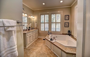 A spacious bathroom with a large bathtub, double vanity with a mirror, and a window with blinds. - The Good House B&B (Natchitoches)