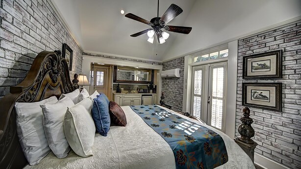 A bedroom with a bed, a ceiling fan, a brick wall, and a kitchen in the background. - The Good House B&B (Natchitoches)
