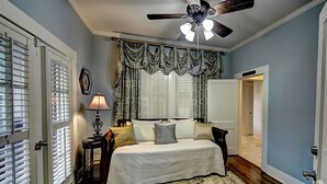 A bedroom with a bed, a ceiling fan, a nightstand, a lamp, and a window with curtains. - The Good House B&B (Natchitoches)