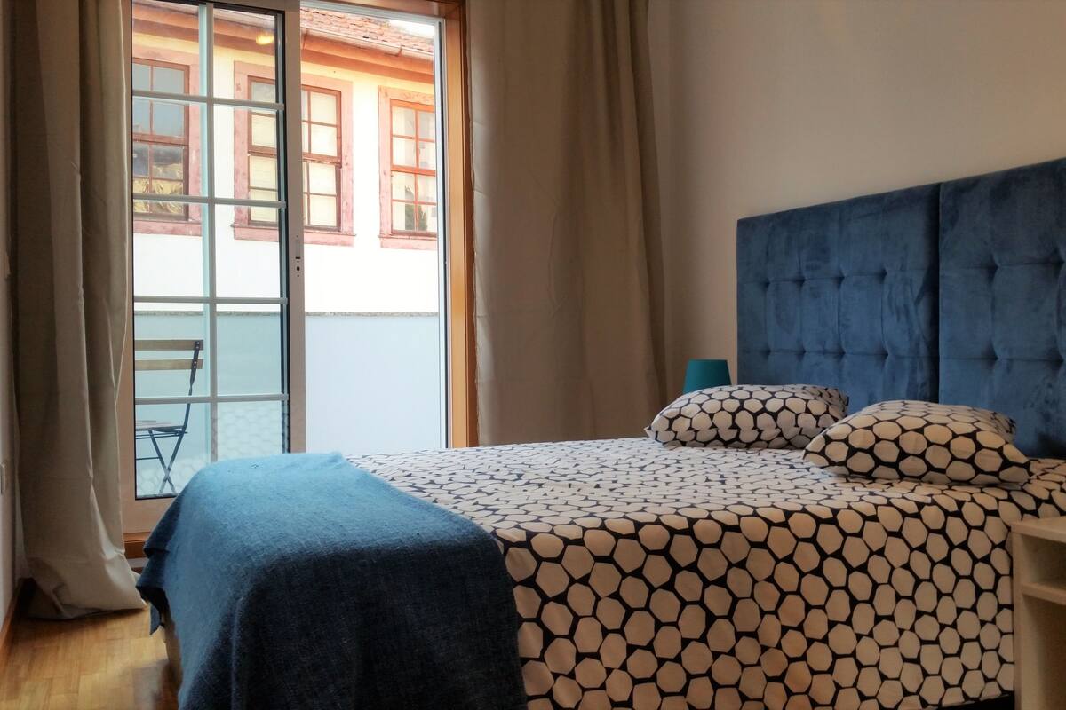 Un dormitorio con cama, mesita de noche, ventana con cortinas y vistas al exterior.