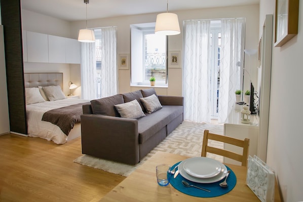 A modern bedroom with a bed, a sofa, a dining table with plates and cutlery, and a window with curtains.