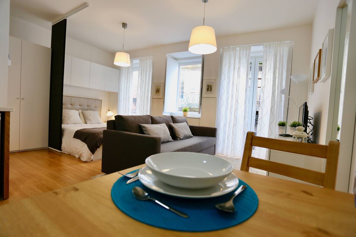Una sala moderna con un sofá, una mesa de comedor con platos y cubiertos, y una cama con cabecera.