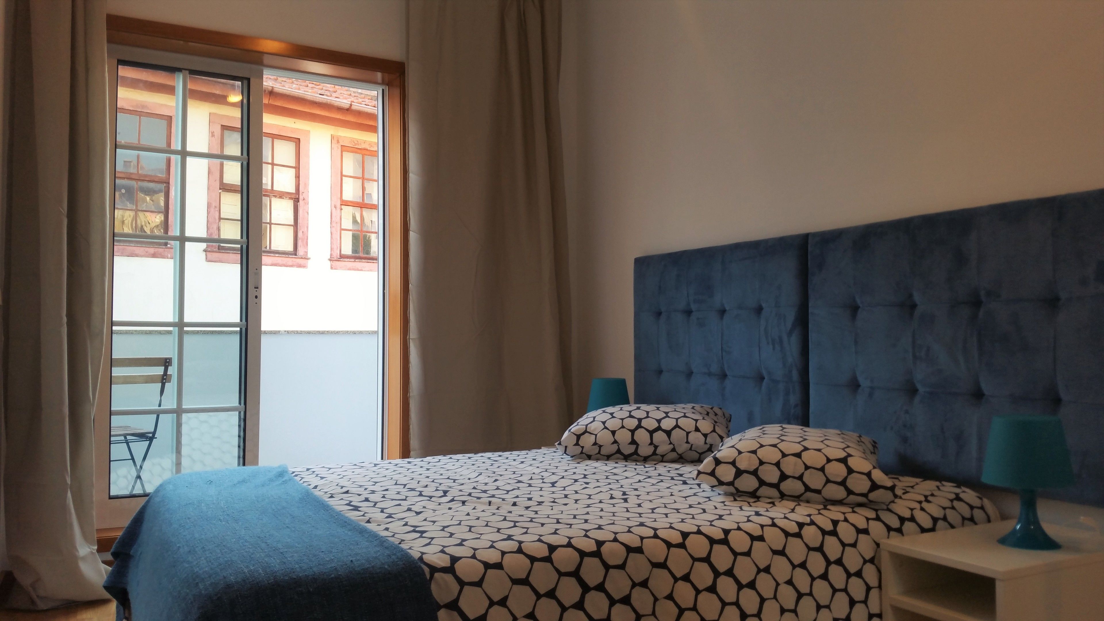 Habitación de hotel con cama, cabecera, mesita de noche, lámpara y vista al exterior a través de una ventana.