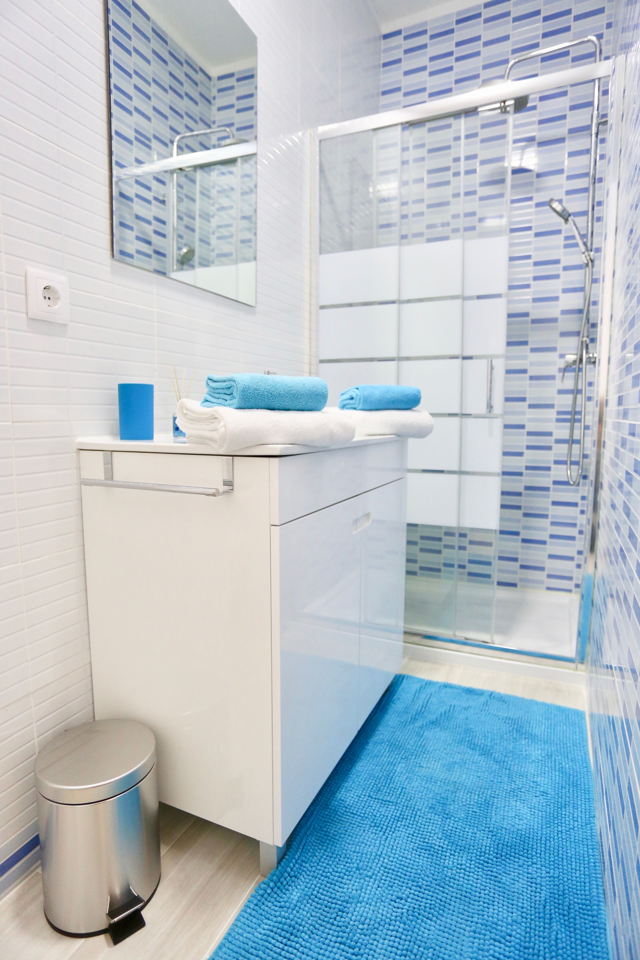 Un baño moderno con un lavamanos blanco, toallas azules, una cabina de ducha de vidrio y un tapete de baño azul.