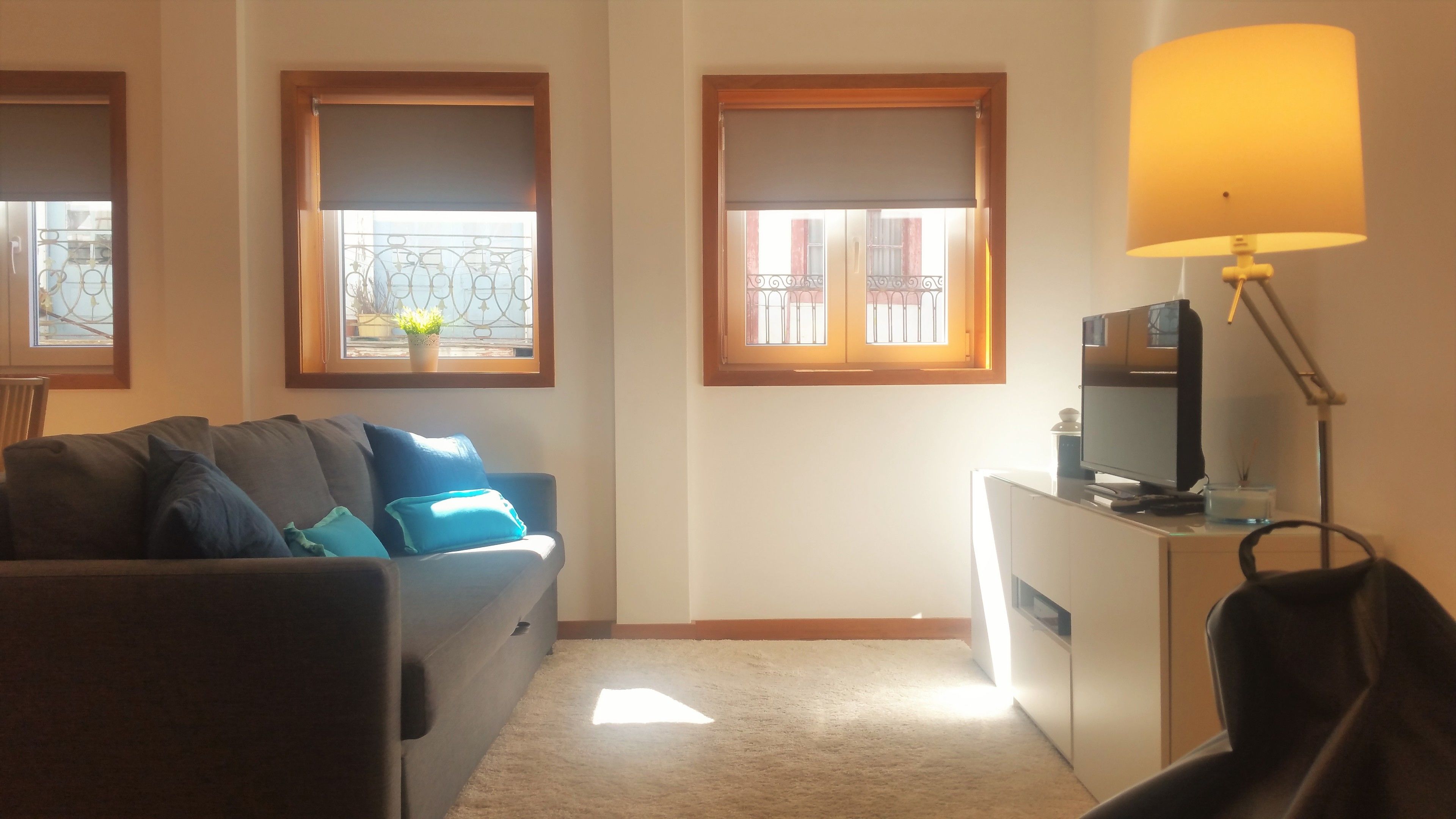 A living room with a sofa, a TV on a stand, and a lamp. There are windows with blinds and a view of the outside.