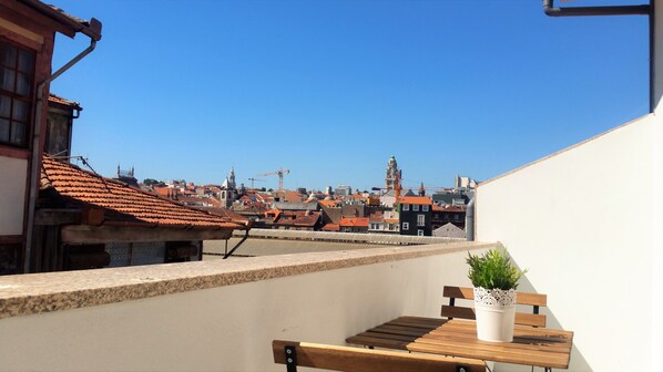 Terraza en la azotea con un banco de madera y una planta en maceta, con vistas a un paisaje urbano de tejados rojos y una torre destacada.
