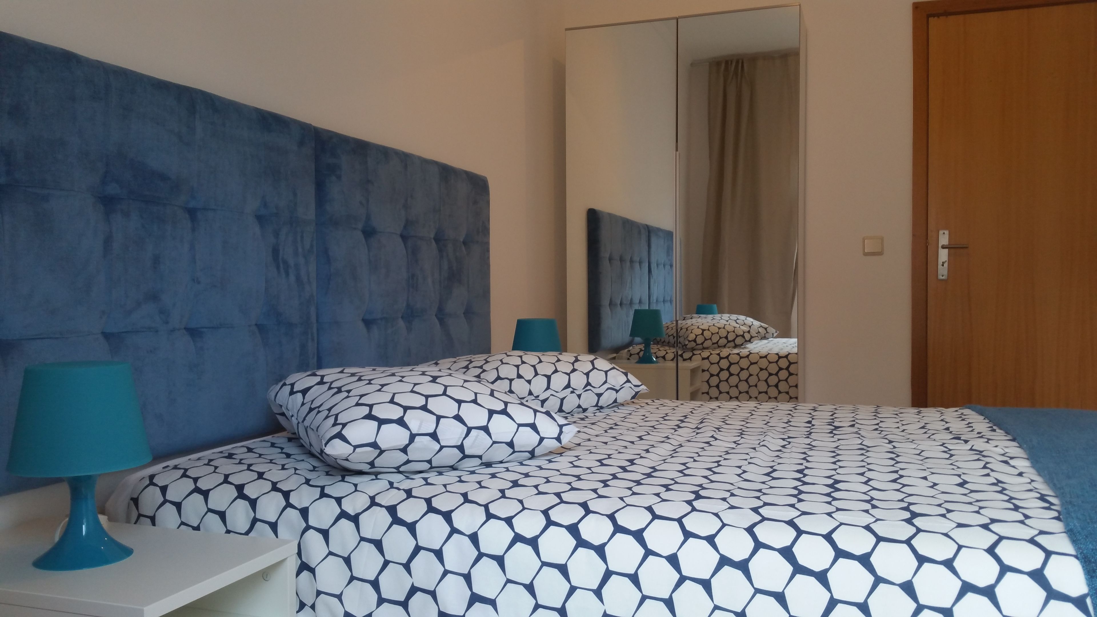 Un dormitorio con una cama grande, un espejo, una puerta de madera y una mesita de noche con lámpara.