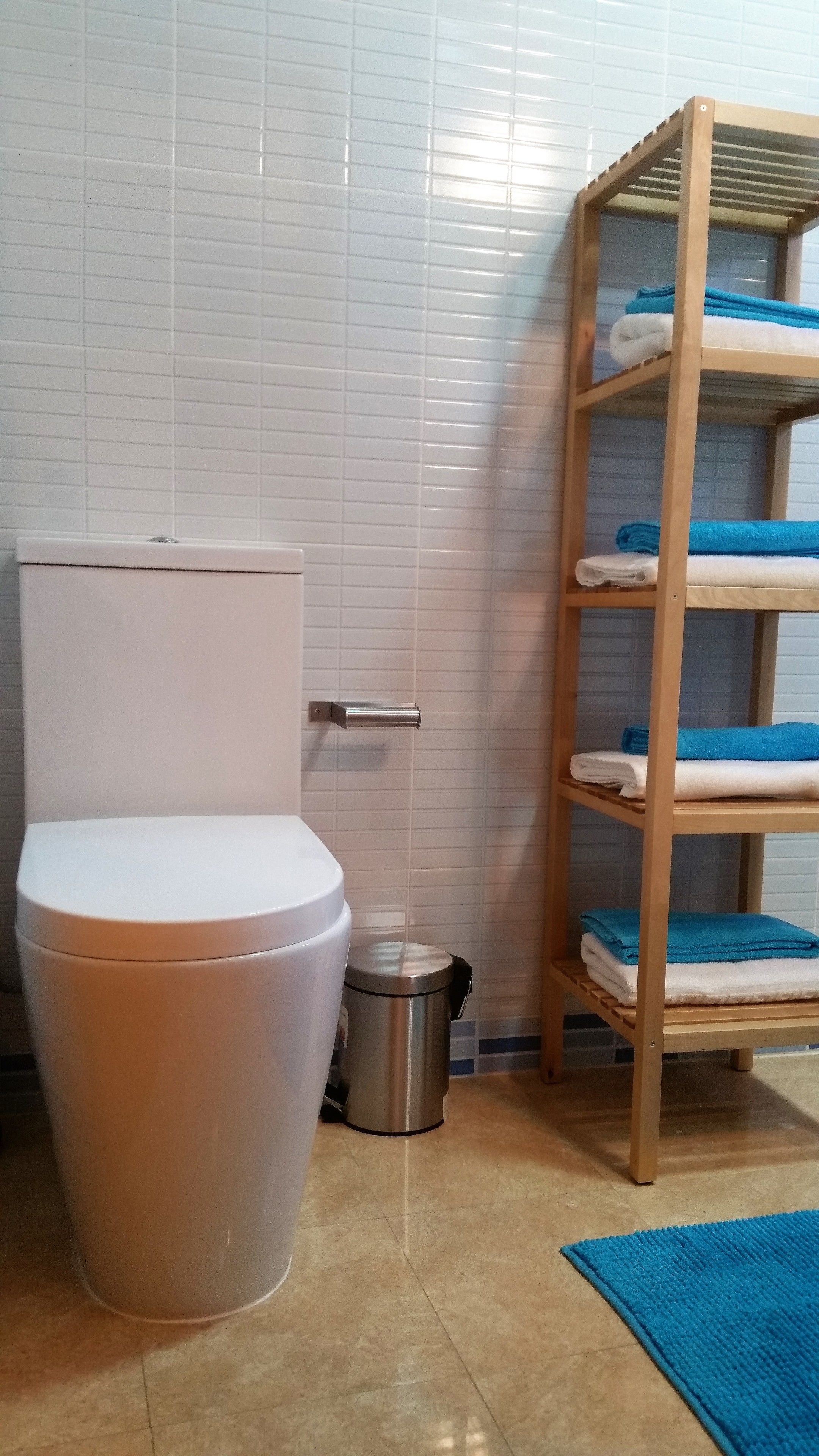 Une salle de bain avec des toilettes, une poubelle et une étagère en bois où sont posées des serviettes.