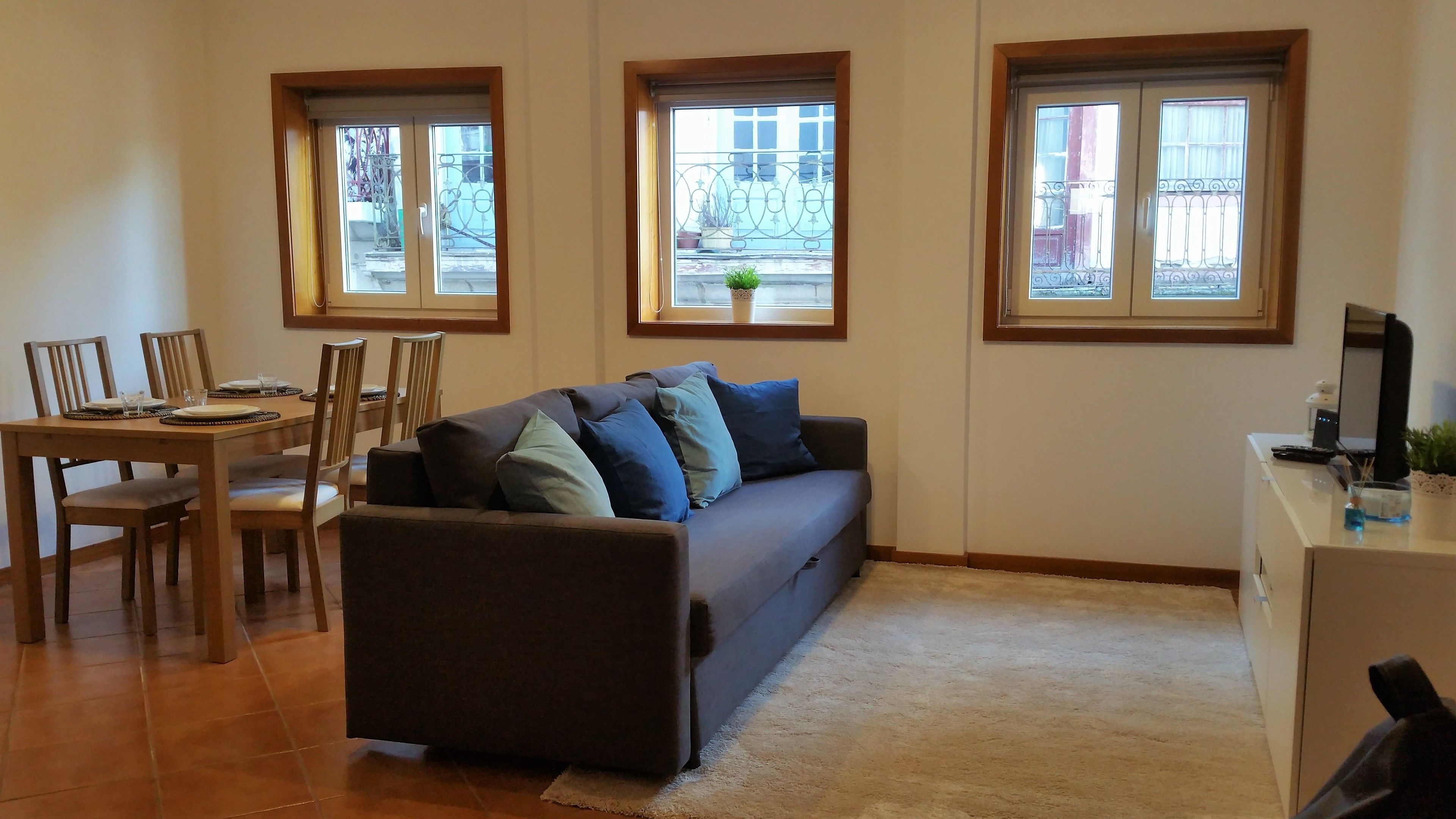 Ein Wohnzimmer mit einer Couch, Sesseln, einem Esstisch und einem kleinen Schreibtisch mit einem Computer. Drei Fenster sorgen für natürliches Licht.