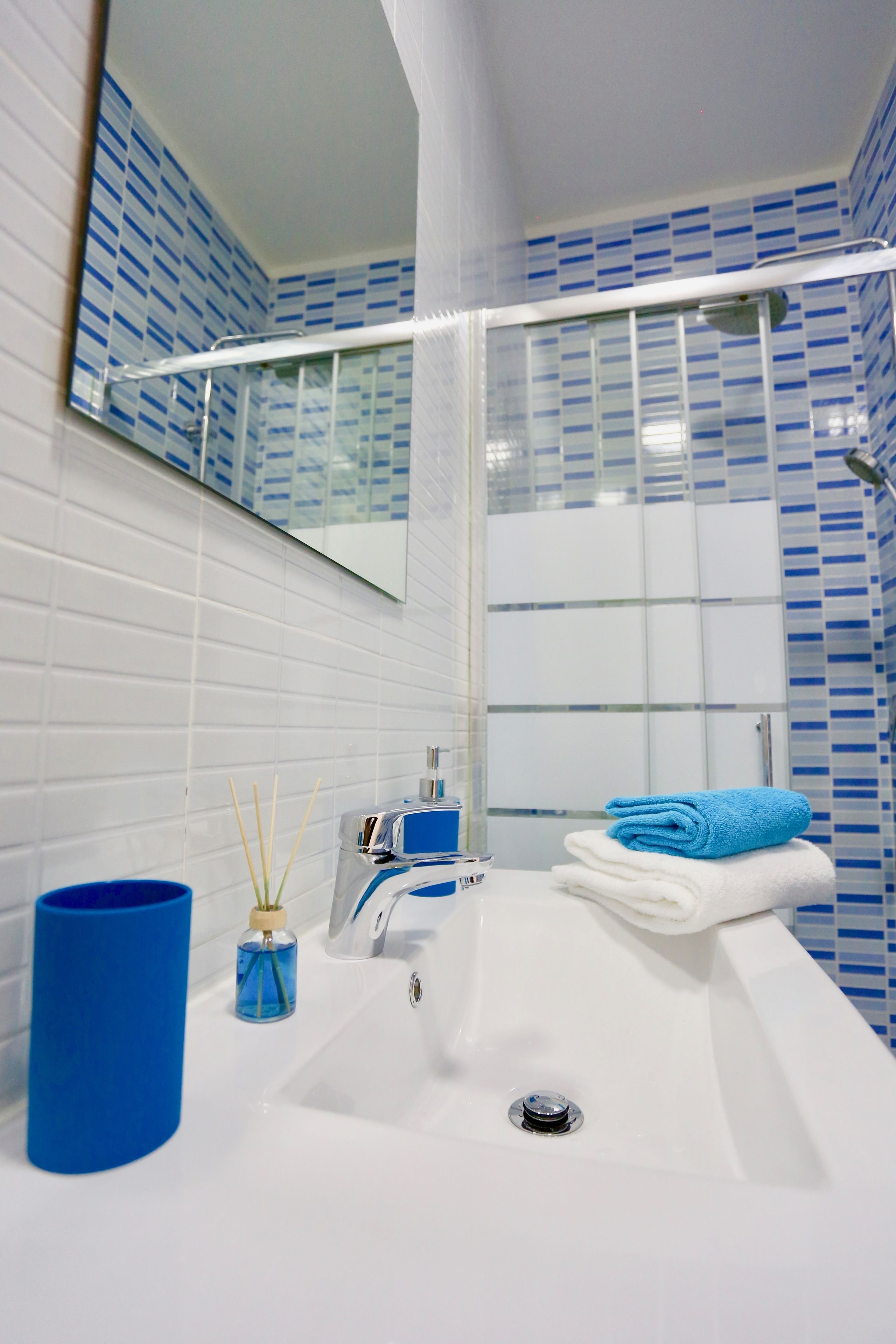 A bathroom with a white sink, blue and white tiled walls, a glass shower enclosure, and a blue candle holder.