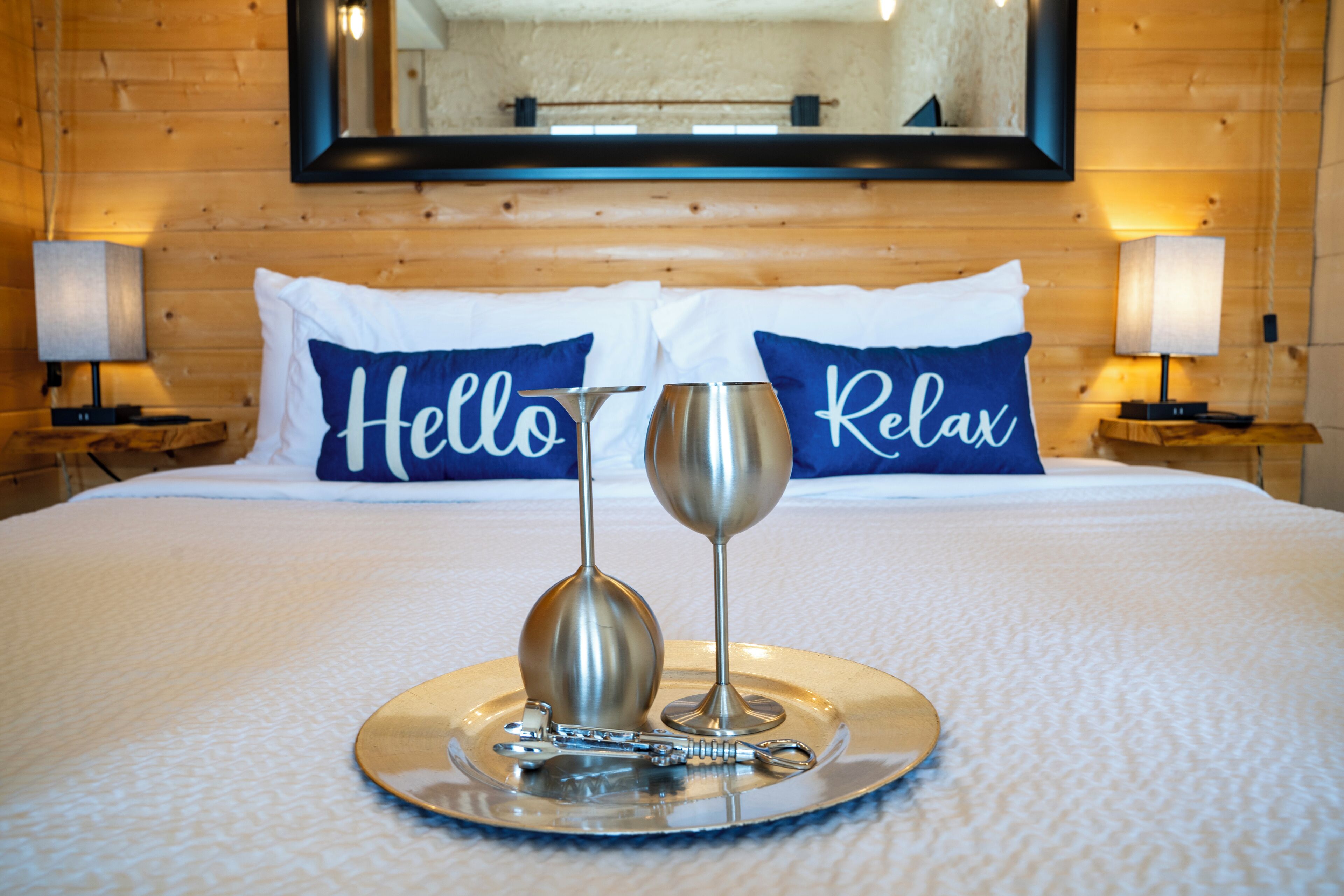 A neatly made bed with two pillows labeled "Hello" and "Relax," a tray with a wine glass and a bottle, and a wooden wall with a mirror above the bed.