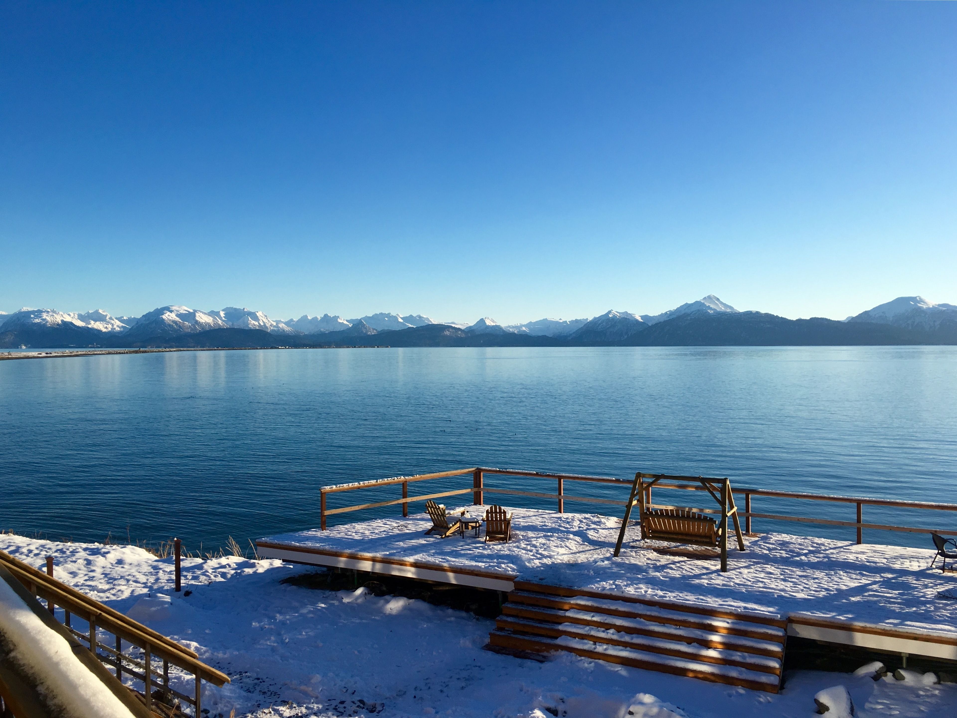 Une terrasse enneigée, dotée de bancs en bois et d’une balustrade, offre une vue sur un lac paisible, avec des montagnes en toile de fond.