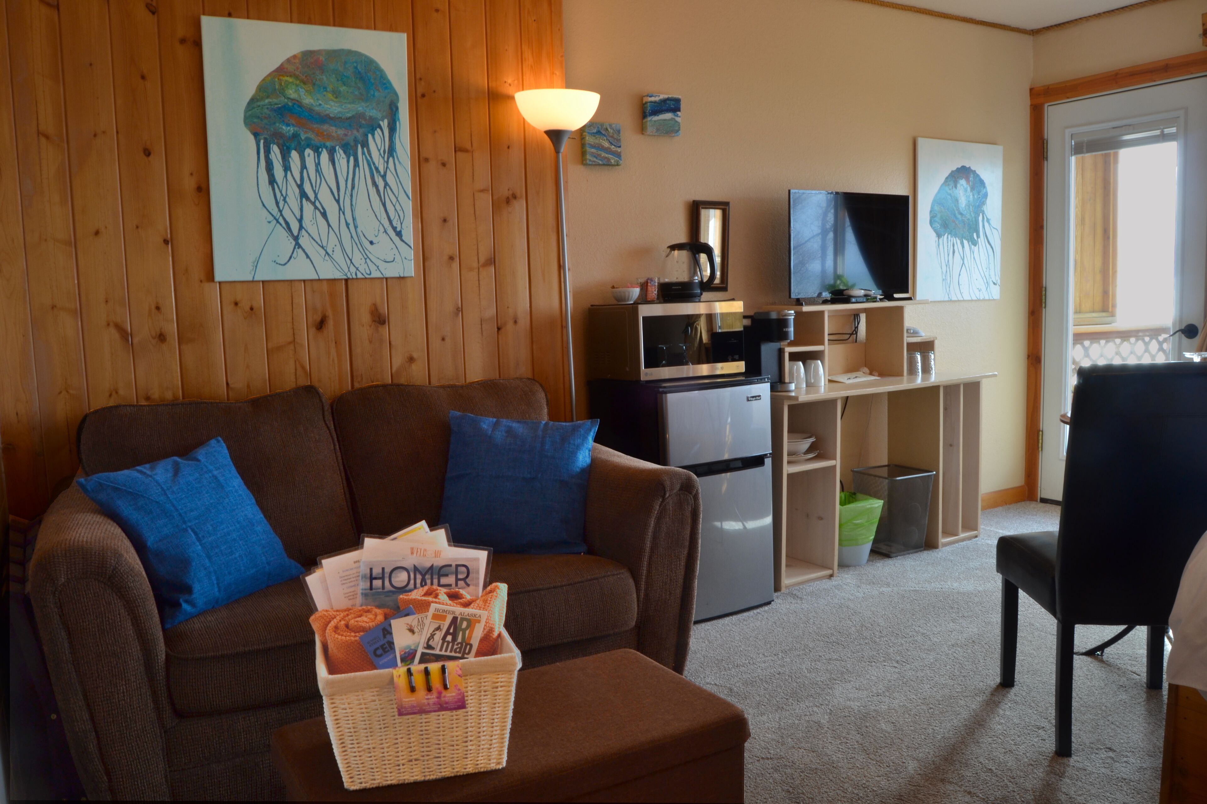 A cozy living room with a brown sofa, blue cushions, a basket of magazines, and a flat-screen TV on a wooden wall.