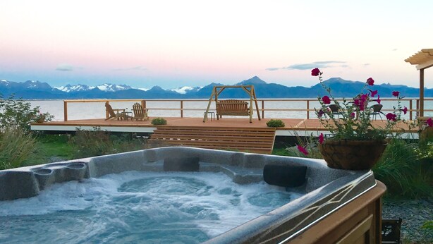 A hot tub with a wooden deck, chairs, and a view of mountains and water. - Homer Inn and Spa (Homer)