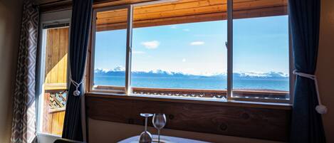 A room with a large window offering a view of a body of water and mountains. There is a bed with a white bedspread and a small table with two wine glasses.