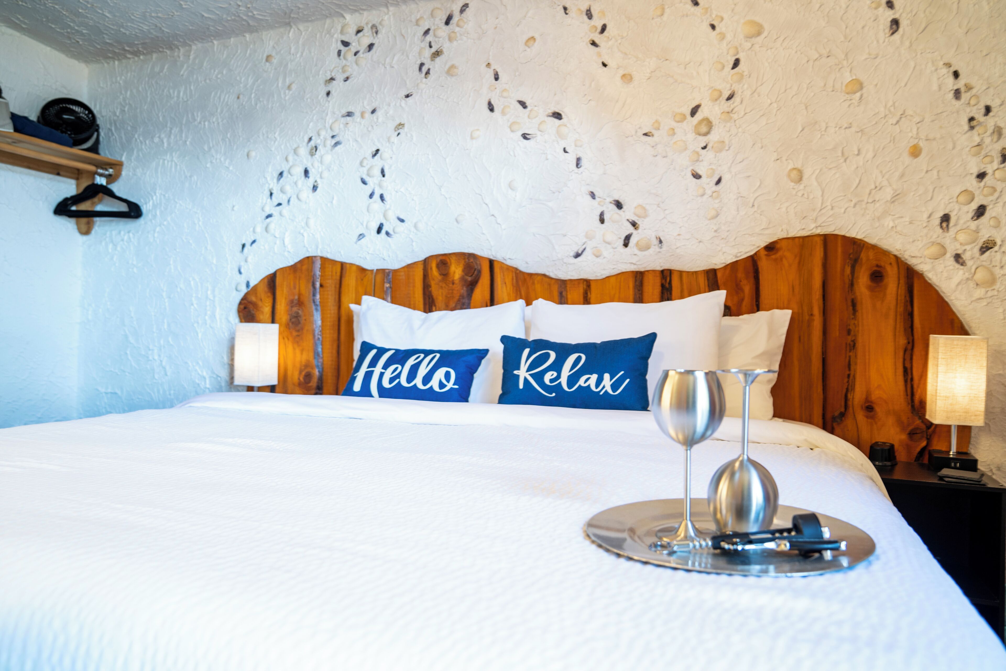 A neatly made bed with a wooden headboard, two pillows with "Hello" and "Relax" text, and a tray with a wine glass and remote control.