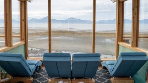 A room with large windows overlooking a beach and mountains. There are three blue chairs and a small table. - Homer Inn and Spa (Homer)