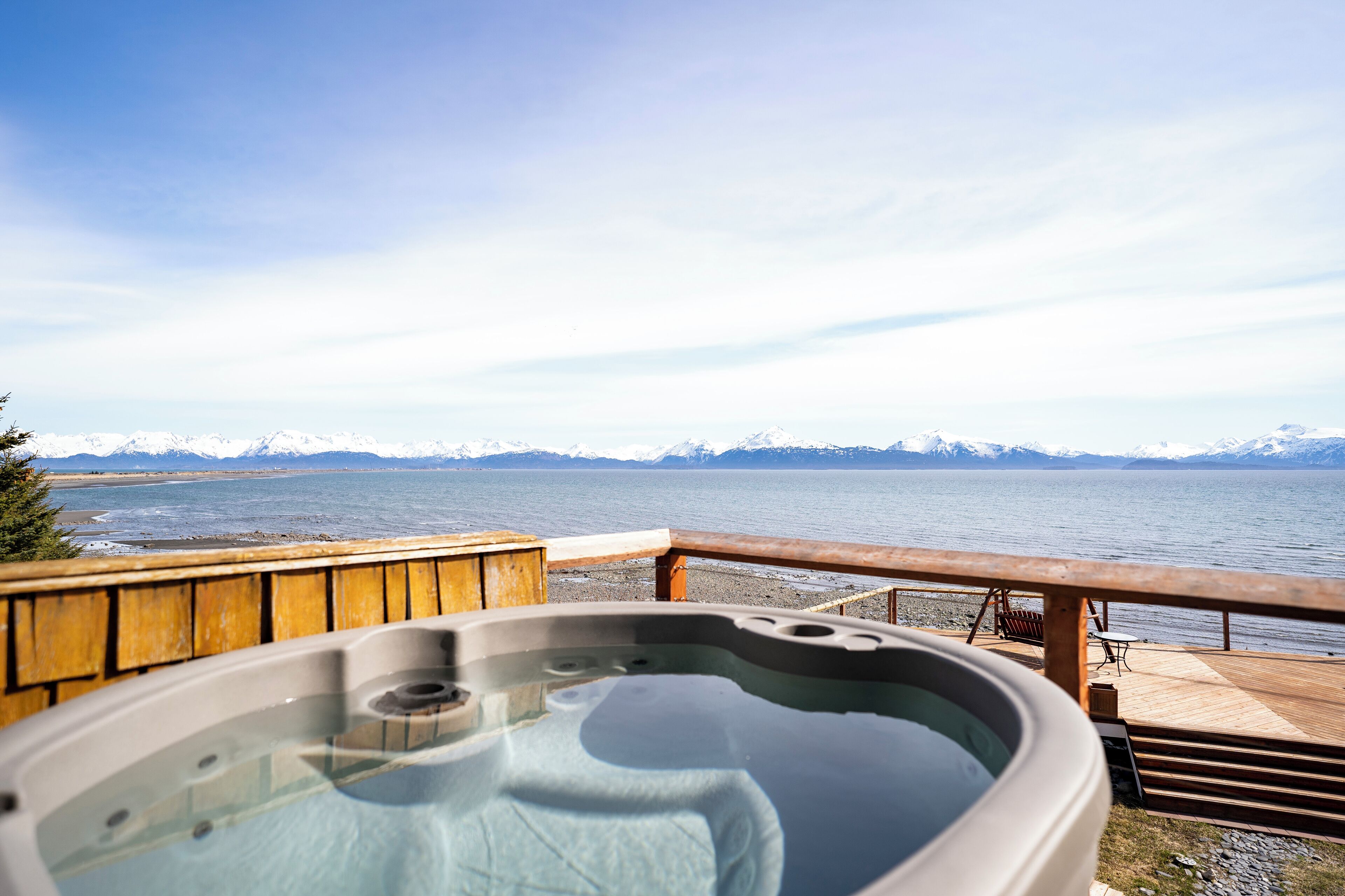 A hot tub on a wooden deck overlooking a lake with mountains in the distance.