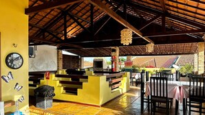 A restaurant with a rustic interior, featuring a wooden ceiling, a brick wall, and a tiled floor. There is a counter with stools and a fire extinguisher visible. - Pousada Alternativa (Aracati)
