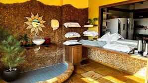 A bathroom with a stone wall, a sun-shaped decoration, a sink, a mirror, and a shower area. - Pousada Alternativa (Aracati)