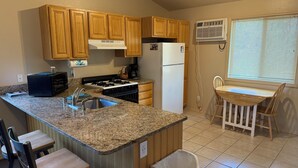 Una cocina con encimeras de granito, gabinetes de madera, estufa a gas, refrigerador, fregadero, basurero y comedor con mesa y sillas.