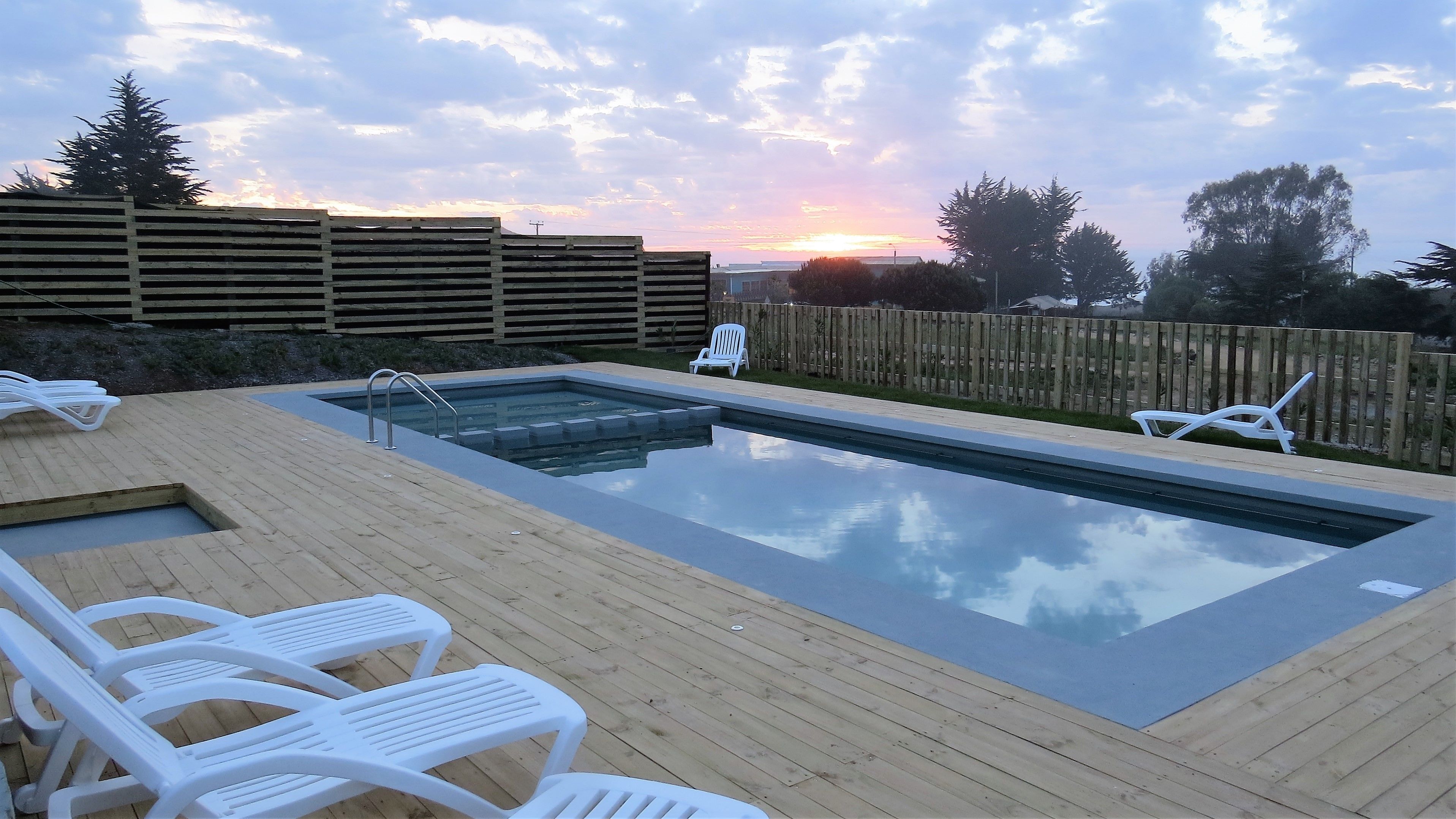 A rectangular swimming pool with a wooden deck and white lounge chairs.