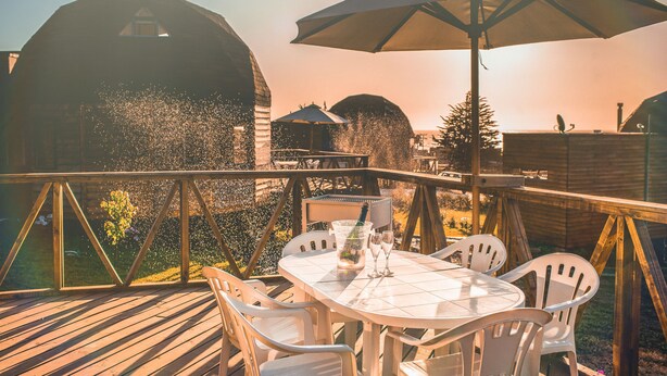 A wooden deck with a table and chairs, a large umbrella, and a view of barns in the background. - Pichilemu Domos (Pichilemu)