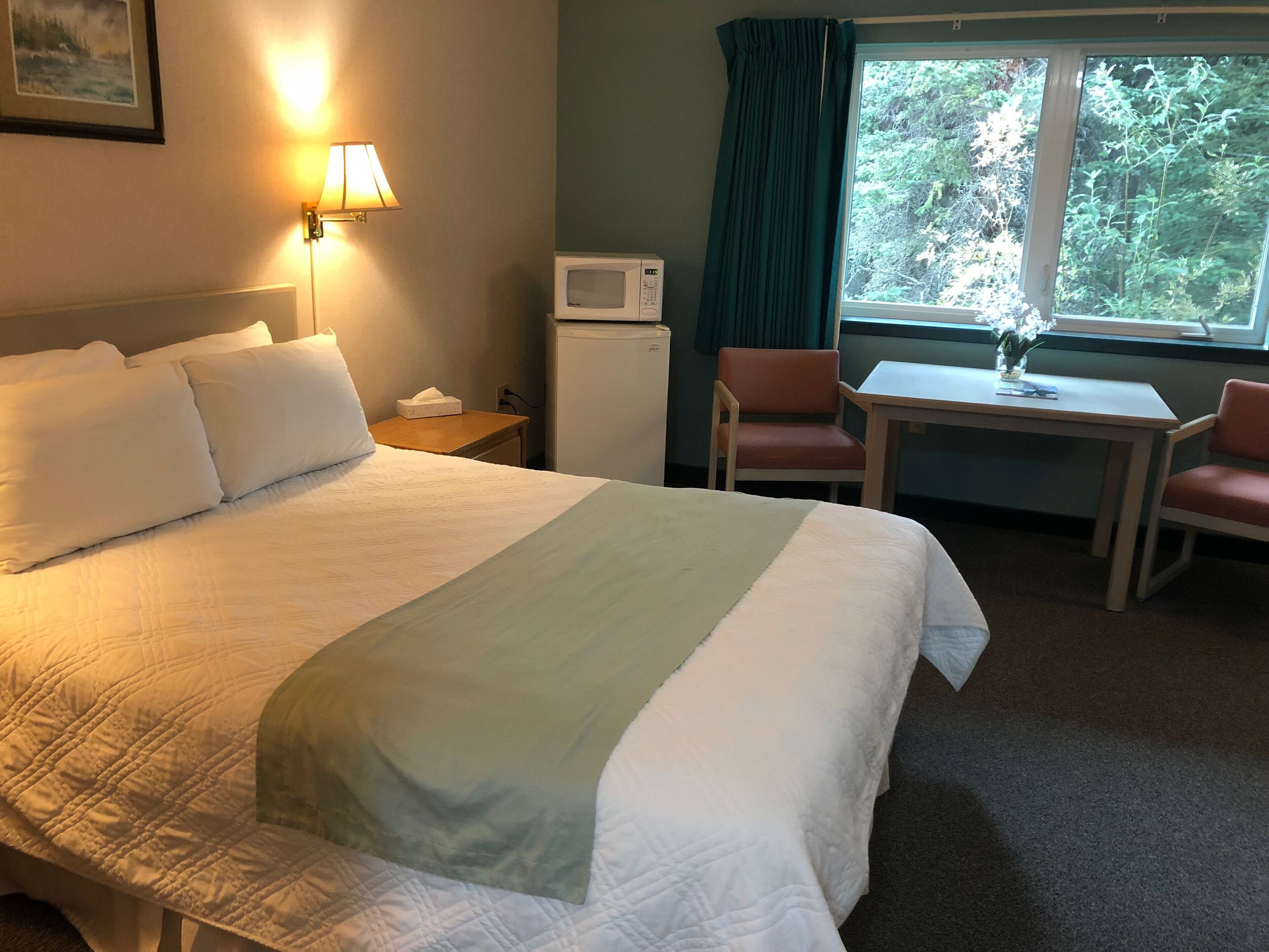 Une chambre d’hôtel avec un grand lit, un bureau, deux chaises, un four à micro-ondes et une fenêtre donnant sur des arbres.